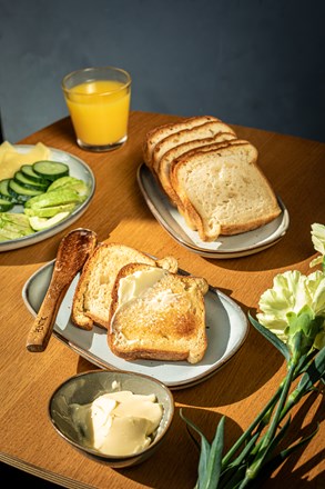 Fazer Gluteeniton Vaalea Paahto -leipä pöydällä kattauksessa, jossa on myös appelsiinimehua ja vihreitä kasviksia.