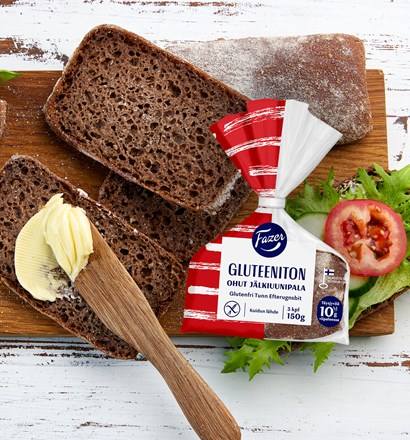 Valkoisella, rustiikkisella tasolla leikkuulauta, jonka päällä Fazer Gluteeniton Ohut Jälkiuunipala -leipiä sekä ilman voita, voilla että erilaisilla leivänpäällisillä (mm. kurkkua, salaattia ja tomaattia).