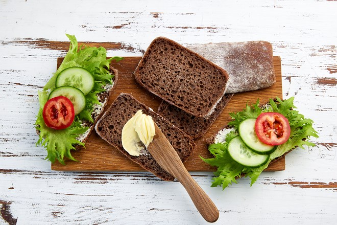 Valkoisella, rustiikkisella tasolla leikkuulauta, jonka päällä Fazer Gluteeniton Ohut Jälkiuunipala -leipiä sekä ilman voita, voilla että erilaisilla leivänpäällisillä (mm. kurkkua, salaattia ja tomaattia).